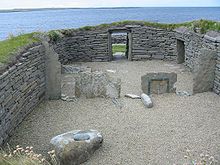A small area of gravel is enclosed by a stone wall built into the surrounding grassy fields. Various large stones sit inside this enclosure and a low doorway has been constructed at the far end. Beyond the doorway, there is a rocky foreshore and a body of water.