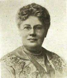 B&W portrait photo of a woman with her hair in an up-do, wearing a white pearl necklace and a patterned jacket.