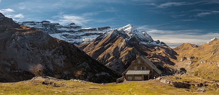 Un petit chalet de pierre à toit pointu dans une montagne brune et rocheuse, qui se couvre de neige en devenant verticale.