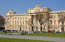 kolorowe zdjęcie. Na pierwszym planie zielony skwer. Za drzewami na dalszym planie dominuje żółta fasada budynku Sejmu Krajowego (współcześnie Uniwersytet im. Iwana Franki). Wejście frontowe bogato zdobione. Pierwsza kondygnacja to łuki, na drugiej kondygnacji dominują kolumny. Wejście na dachu budynku zdobią rzeźby i flaga Ukrainy. Flaga jest w kolorach niebieskim i żółtym.