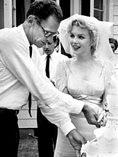 Foto recortada de Monroe y Miller cortando el pastel en su boda. Su velo está levantado de su rostro y él lleva una camisa blanca con una corbata oscura.