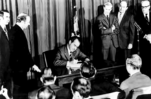 Photograph showing President Nixon seated at a desk signing the bill, with multiple people watching him; those to his right and left are standing, and those facing him in the foreground are seated