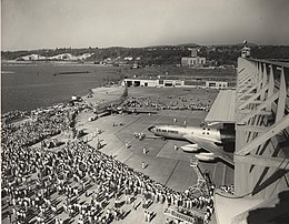 Une foule de personnes forme un arc de cercle autour de deux avions : un à hélices, sur la gauche, en vue de trois-quarts avant, et un sur la droite, sortant d'un hangar, dont est visible le côté gauche ; une estrade est installée à proximité.