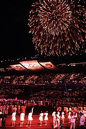 Photographie de l'intérieur d'un stade, de nuit. Des athètes sont regroupés sur le terrain central, et dans le ciel est tiré un feu d'artifice.