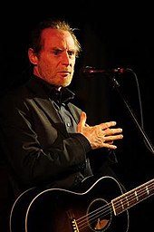 An older man wearing a black shirt, holding a guitar
