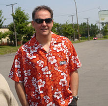 Photographie d'un homme portant des lunettes noires et une chemise rouge à fleurs blanches.