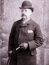 Man with a moustache wearing a bowler hat, topcoat, and leather gloves. He holds a cane in the left hand, and a cigar in the right.