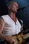 A man with a v-neck, white t-shirt wearing a necklace and bracelet standing behind a microphone, holding a guitar.