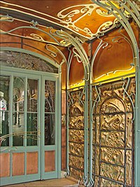 Photo en couleur d'un coin de pièce aux tons chauds dont les murs en relief tarabiscotés sont encadrés d'arabesques en fer forgé