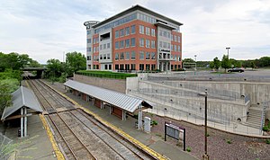 A railway station in a cut, with a switchback ramp leading from one platform to a parking lot next to a commercial building