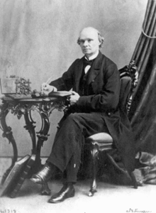 Photo of fair-skinned man, balding, wearing a mid-Victorian business suit and seated at a table