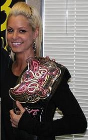 A blonde Caucasian woman holding a wrestling championship on her left shoulder. She is wearing a black, long-sleeve t-shirt and long silver earrings. The championship belt has a black strap, and the front of the belt is in a pink butterfly design, with the word "Divas" in white.