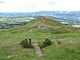 The southern end of the foundations of a small rectangular chapel on a mountain ridge. The entrance to the chapel is marked by two stones. Beound the stones the ridge falls away gently. In the distance is a green flat valley.