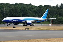 Side view of aircraft on runway. Runway surfaces in foreground and forest in background.