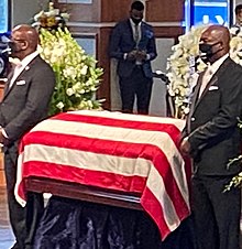 Two men wearing dark business suits stand on either side of a casket.  The casket has been draped with a flag, a sign of military or government service in that country