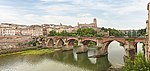 A distant view of a small city in the background and a narrow bridge hidden by a few trees along a river.