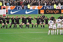 Equipo de rugby vestio todo de negro, mirando al camara, con el maga rodilla doblao y mirando hacia un equipo vestio de blanco