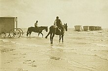 2 chevaux montés, dont 1 tire une cabine cubique à 4 roues vers une rangée de cabines semblables déjà placées au large de la plage.