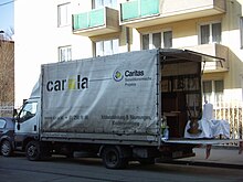 LKW mit der Aufschrift „carla“ (Caritas Lager) parkt mit heruntergelassener Hebebühne vor einem Wohnhaus.