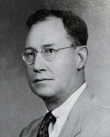 A black-and-white photographic portrait of a middle-aged man from the shoulders up, wearing a suit, with a necktie, and eyeglasses