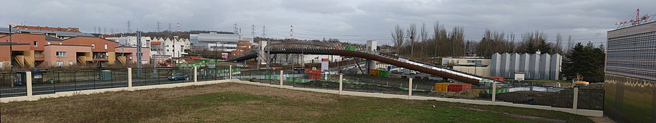 Le premier ouvrage d'art construit sur le Tram Express Nord : la passerelle de Villetaneuse-Université, photographiée pendant son chantier de construction, desservant la gare construite dans un second temps à cheval sur les voies, et qui relie l'Université Paris-XIII, dont on voit le gymnase universitaire à droite du cliché, et le quartier du Centre-ville, à gauche. Au fond à droite, le Centre nautique Jacques-Duclos, démolit depuis.