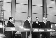 Cuatro hombres están detrás de podios con sus nombres de países de Francia, Alemania, Reino Unido y Estados Unidos, frente a un telón de fondo de la Torre Eiffel