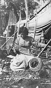 Foto in bianco e nero di una donna inginocchiata a terra mentre fabbrica una racchetta da neve. Indossa una camicia a scacchi e un vestito bianco che resta chiuso sulla racchetta da neve. Intorno a lei vi sono quattro telai di racchette da neve da fabbricare appoggiate su un tipi.