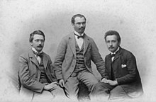 Three young men in suits with high white collars and bow ties, sitting.