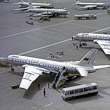 tarmac d'un aéroport. au premier plan, des passagers embarquent dans un Tu-104, depuis un bus. en arrière plan, un autre tu104 et un il-18 à hélices.