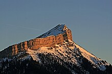 Sommet enneigé présentant un pan incliné incurvé vers la gauche et une paroi abrupte reflétant le soleil à droite.