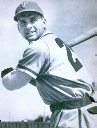 A man in a white baseball uniform and a dark cap holds his bat, ready to swing