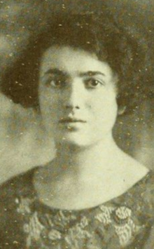 A young woman with fair skin and dark hair cut in a wavy bob; she is wearing a dress or top with a scooped neckline and embroidered flower designs