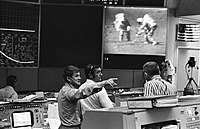 A control room; visible on a large screen are two astronauts walking on the Moon