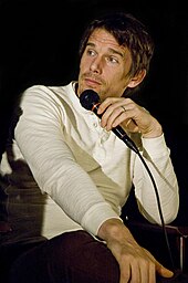 A Caucasian male with light brown hair and light brown stubble is facing to the left. He is wearing a white long sleeve shirt, and is holding a black microphone.