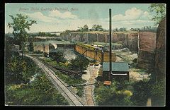 A 1911 postcard showing a quarrying operation in Portland Connecticut with cliffs of brownstone in the background, rail lines for loading and transporting stone, industrial buildings, rail carts, and other parts of the operation.