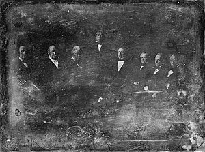 Photographie très endommagée montrant Taylor debout entouré de sept hommes assis.