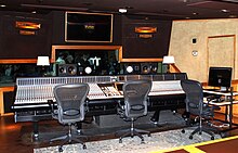 A black room containing video screens, a large mixing desk and three swivel chairs.