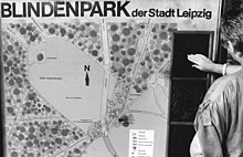 Das Schwarzweißfoto von 1986 zeigt eine senkrecht aufgestellte Informationstafel. Unter der in Schwarzschrift ausgeführten Überschrift „BLINDENPARK der Stadt Leipzig“ zeigt der größte Teil der Tafel einen gezeichneten Plan der Parkanlage mit einer großen Wiese, Wegen und bewaldeten Flächen. Im rechten Fünftel der Tafel sind senkrecht untereinander mehrere schwarze Reliefs angeordnet, die offenbar Ausschnitte des Plans tastbar darstellen. Am rechten Bildrand steht eine Frau, die das oberste dieser Reliefs mit ihren Fingerspitzen betastet.