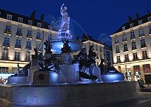 La fontaine, de nuit, éclairée en bleu sur fond d'immeubles éclairés en blanc.