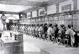 Photographie d'une très grande salle de travail ; un mur est intégralement occupé par des appareils téléphoniques ; une vingtaine de femmes assises et visibles de dos y travaillent.
