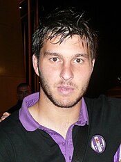 The upper body of a man in a red coat. He has black hair and is holding a large silver trophy.