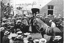 Photo en noir et blanc présentant une cour où des hommes et des femmes en chapeaux entourent un cheval.