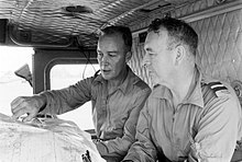 Black and white photograph of two men wearing military uniforms seated in the cabin of a vehicle. The man on the left is pointing at a location on a map.