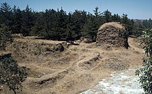 Ruïnes cobertes d'herba i d'arbusts amb un fons de boscs de pi baix. Hi ha runes d'una torre quadrada a la dreta, tot el que queda del Temple de Tohil, amb les restes de les parets del camp de joc de pilota a l'esquerra al primer plànol.