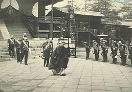 Photo de l'empereur du Japon en train de sortir d'un temple. Un prêtre shinto le précède, et il est suivi par six hommes en uniformes de cérémonie de hauts gradés militaires. Le long de l'allée, un groupe d'hommes s'inclinent à son passage.