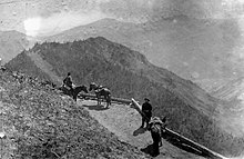 Vue depuis un sommet d'un chemin en contre-bas, où l'on observe deux chevaux et un cavalier qui attend. Le cavalier manquant est parti poser son chapeau à l'autre extrémité du passage risqué, afin de signaler que la voie étroite est occupée. En arrière-plan, un vide se révèle, montrant que la scène est à flanc de falaise.