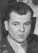 Clean-shaven Caucasian man wearing a suit and tie. He is glancing to the side and has his wavy hair combed back.