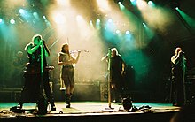 Four singers perform in the spotlights on stage. One is a female in shorts, a gauzy beige skirt and a leather belt, and plays the fiddle, The males are wearing black sleeveless shirts, black pants and have military-looking gear. A rock band is in the background.