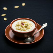 A image of Chhena kheeri in a rich white color with yellow hints and some nuts on top of it. The background is in a distinct black color with many nuts in the background, and the bowl holding it is in a a brown color. There is a spoon on the side of the bowl, which is made with steel, has engraved designs on the end tip of it.