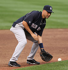 A man in a blue baseball uniform with "New York" written on the front in grey letters and a navy hat with white letters "N" and "Y" interlocking prepares to catch a ground ball with his baseball glove.